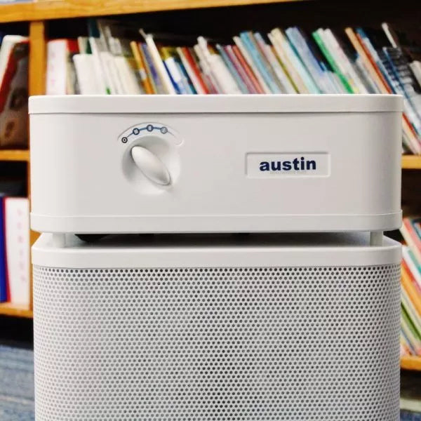 A white Austin Air Healthmate Plus Air Purifier with a control dial sits in front of a bookshelf filled with colorful books.