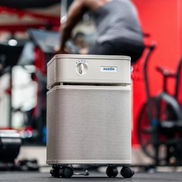 A silver Austin Air Healthmate Plus Air Purifier on wheels sits in the foreground of a gym, with blurred exercise equipment and a person working out in the background.