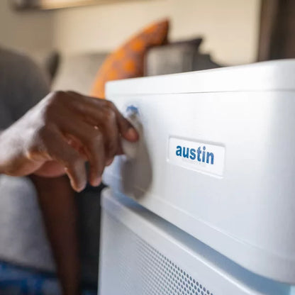A person adjusts the knob on a white Austin Air Healthmate Plus Air Purifier labeled “austin,” with a softly blurred couch and cushion visible in the background.
