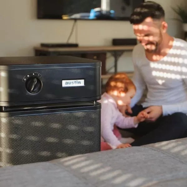 A black Austin Air Healthmate Plus Air Purifier stands in the foreground as a man and child play together on the sunlit floor in the background.