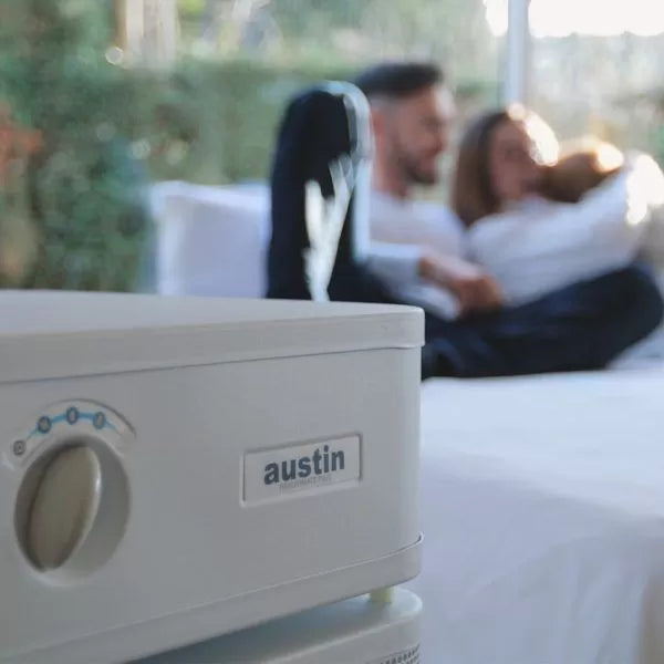 An Austin Air Healthmate Plus Air Purifier is in focus in the foreground, with a blurred family sitting together on a bed by a window with greenery outside.