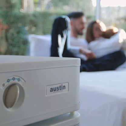 An Austin Air Healthmate Plus Air Purifier is in focus in the foreground, with a blurred family sitting together on a bed by a window with greenery outside.