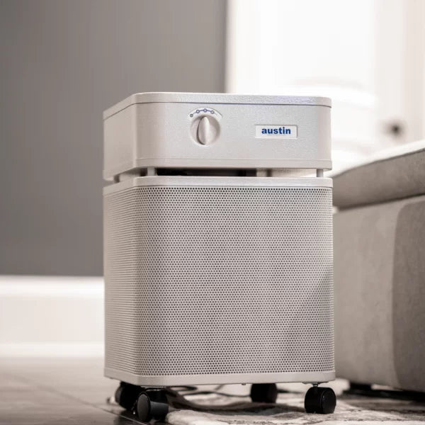 A silver Austin Air Healthmate Air Purifier with a control knob and small wheels is placed on the floor beside a light gray sofa in a modern room.