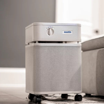 A silver Austin Air Healthmate Air Purifier with a control knob and small wheels is placed on the floor beside a light gray sofa in a modern room.