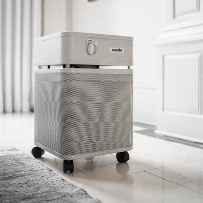 An Austin Air Healthmate Air Purifier with a control knob, perforated metal body, and wheels stands on a tiled floor beside a rug in a bright, modern room with white walls and curtains.