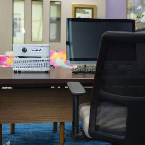 A modern office desk with a computer monitor, mesh-back chair, and an Austin “it” Air Purifier; a colorful sofa and vibrant wall art brighten the background.