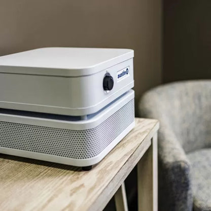 An Austin “it” Air Purifier sits on a light wooden table beside a gray upholstered chair in a softly lit room with beige walls.