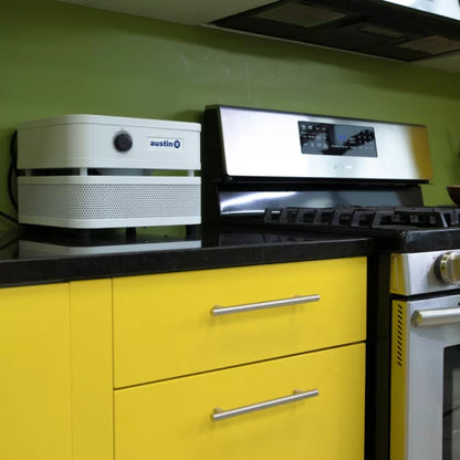A kitchen features yellow cabinets, a black countertop, a stainless steel stove, and an Austin “it” Air Purifier on the counter beside the stove. The green wall in the background adds a pop of color.